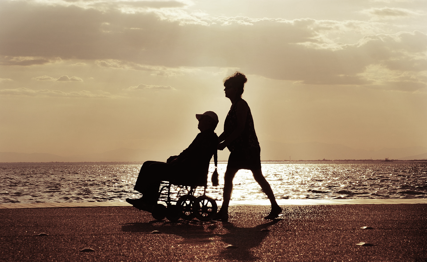 Äldre man i rullstol och en kvinna bakom, gåendes på en badstrand 